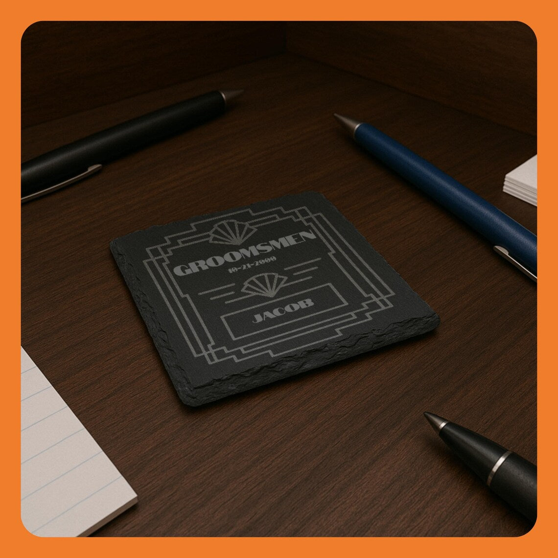 A black coaster with 'Groomsmen' and 'Sachets' engraved, placed on a wooden desk with pens and notepads.